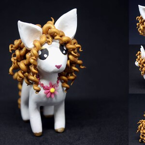 May include: A handcrafted white pony figurine with gold-colored curly hair and a pink flower necklace. The pony has large, expressive eyes and gold-painted hooves. The figurine is displayed against a dark background.
