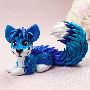 May include: A blue and white furry figurine of a dog with a spotted pattern. The figurine is lying down with its head turned to the side.