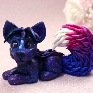 May include: A handmade figurine of a stylized fox-like creature. The figure is primarily dark blue with a galaxy-like effect, speckled with white dots. The tail transitions from white to pink and then to blue. The creature is in a resting pose.