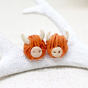 May include: A pair of orange and white Highland Cow earrings. The earrings are made of polymer clay and are about 1 inch in diameter.
