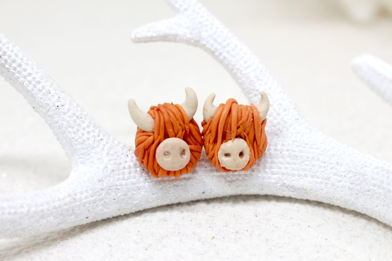 May include: A pair of orange and white Highland Cow earrings. The earrings are made of polymer clay and are about 1 inch in diameter.