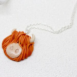 May include: A necklace with a brown and white clay pendant shaped like a Highland cow head. The cow has a fluffy orange mane and white horns.