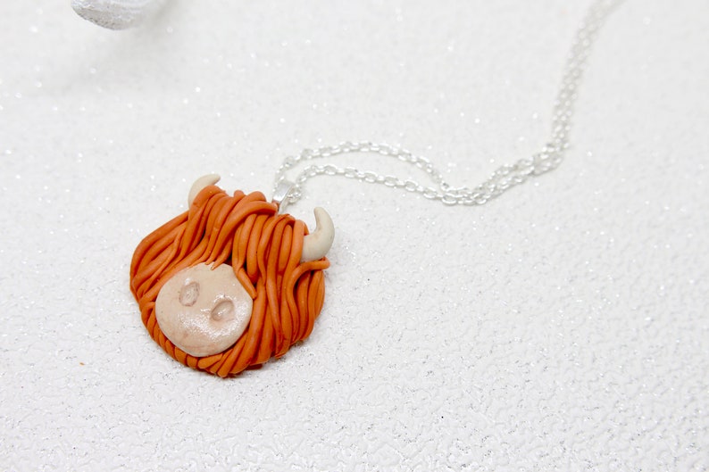May include: A necklace with a brown and white clay pendant shaped like a Highland cow head. The cow has a fluffy orange mane and white horns.