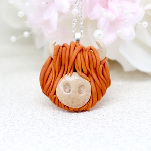 May include: A handmade necklace featuring a brown and white clay Highland cow head pendant. The pendant is attached to a silver chain.
