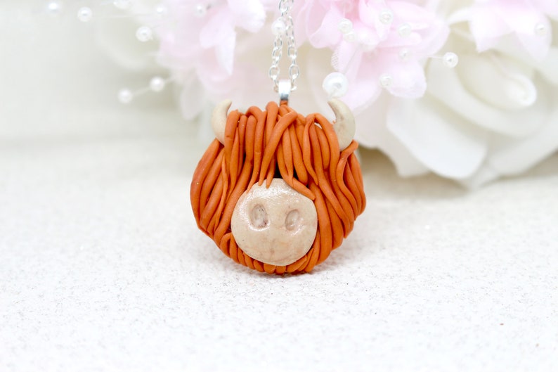 May include: A handmade necklace featuring a brown and white clay Highland cow head pendant. The pendant is attached to a silver chain.