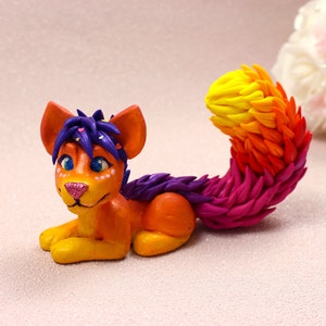 May include: A handmade orange and yellow figurine of a stylized feline creature with a rainbow tail. The figurine has purple hair and blue eyes. The tail transitions from yellow to pink. The creature is lying down.