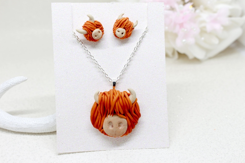 May include: A set of Highland Cow earrings and a necklace. The earrings and pendant are made of orange and white polymer clay and feature a Highland Cow head with fluffy hair.