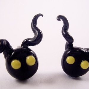 May include: Two black, rounded figurines with yellow eyes and curved, horn-like appendages. The figurines have a glossy finish and are set against a white background. The design is reminiscent of a cartoon character.