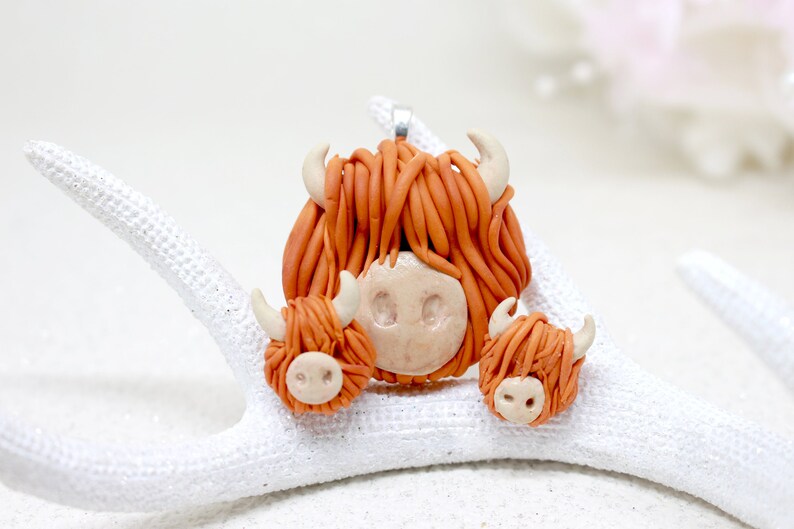 May include: A set of three handmade polymer clay Highland Cow charms. The largest charm is a pendant with a silver loop. The two smaller charms are earrings. All three charms are orange and brown with white faces.