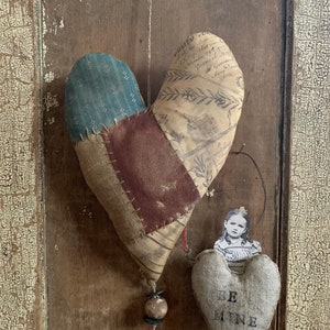 May include: Two fabric hearts, one with a patchwork design and the words "Be Mine" on the other. The larger heart is made of different fabrics in shades of blue, brown, and white. The smaller heart is a light gray color.