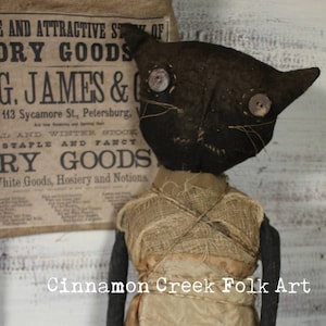 May include: A black rag doll wearing a burlap dress with brown polka dots. The doll has a black cat face with button eyes and a stitched smile. The doll is standing on a wooden surface with a bowl of speckled eggs in the foreground. The text "Cinnamon Creek Folk Art" is visible below the doll.