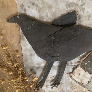 May include: A black crow made of fabric with stitched details. The crow is perched on a white fabric background with brown stains.