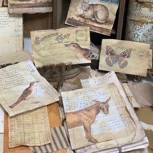May include: A collection of vintage ephemera, including postcards, envelopes, and paper scraps, featuring images of cats, birds, and a donkey. The items are arranged on a wooden surface and have a rustic, aged appearance.