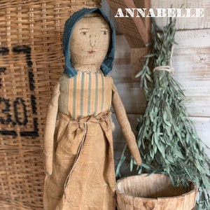 Może przedstawiać: Ręcznie robiona lalka z szmatki o imieniu Annabelle, ubrana w niebieski czepek, żółto-niebieską w paski koszulę i brązową spódnicę. Lalka stoi na drewnianej podstawie i trzyma mały drewniany kubełek. Lalka stoi przed wiklinowym koszem i wiązką suszonych zielonych roślin.