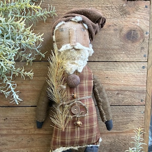 May include: A handcrafted Santa Claus doll with a brown hat and beard. The doll wears a plaid coat with a button closure and a rope belt. It holds a sprig of pine and has blue boots with lace detailing. The doll is set against a wooden background.