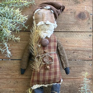 May include: A handmade Santa Claus doll with a brown hat, beard, and plaid vest. The doll has dark blue boots with crisscross laces and is decorated with a sprig of pine. The doll is made of fabric and has a rustic, vintage aesthetic.