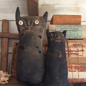 May include: Two black cat dolls with button eyes and stitched facial features. The dolls have long, braided tails and are displayed on a wooden shelf. The dolls are made of a dark, textured fabric and have a rustic, handmade appearance.