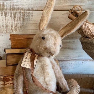 Könnte beinhalten: Ein handgemachtes Plüsch-Hasenspielzeug mit braunen Ohren und einem weißen, flauschigen Körper. Der Hase trägt ein braunes Band mit einem Vintage-Etikett, auf dem "BLACKBERRY TRACTOR" steht, und sitzt auf einer Holzoberfläche.