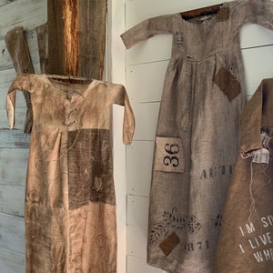 May include: Three vintage-style dresses made from brown fabric with various patches and markings. The dresses are hanging on a white wall with a wooden beam in the background. The dresses have different designs and markings, including a number "36" and the word "AUTUMN".