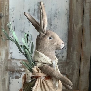 Peut inclure: Un lapin en tissu fait main portant une robe marron et blanche à motif floral vert. Le lapin tient une branche de feuilles vertes dans ses pattes.