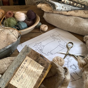 May include: A close-up of a craft project with a variety of yarn balls, fabric scraps, and a needlepoint pattern of a deer. The pattern is on a white sheet of paper and the yarn balls are in a white ceramic bowl. The fabric scraps are in a pile on the table. The text on the paper reads "The Tailoring.  Victoria and Church.  Crick. No. 1."