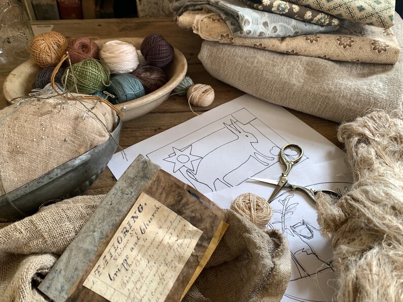 May include: A close-up of a craft project with a variety of yarn balls, fabric scraps, and a needlepoint pattern of a deer. The pattern is on a white sheet of paper and the yarn balls are in a white ceramic bowl. The fabric scraps are in a pile on the table. The text on the paper reads "The Tailoring.  Victoria and Church.  Crick. No. 1."