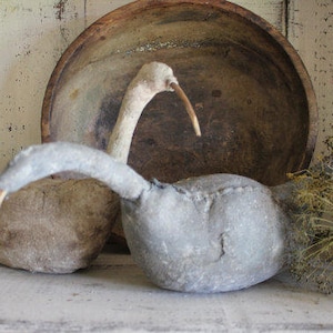 Puede incluir: Dos esculturas de pájaros de tela gris con cuellos largos y picos, sentadas sobre una superficie de madera blanca frente a un cuenco de madera marrón.