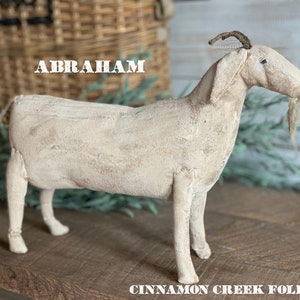 May include: A white, handmade, folk art goat figurine with the name "Abraham" written above it. The goat has a long, thin neck and a long, thin beard. The goat is standing on a wooden surface.