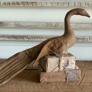 May include: A brown fabric bird with a long neck and tail, perched on top of a stack of three wrapped packages. The packages are tied with twine and have labels with text.