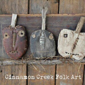 May include: Three rustic folk art faces made of fabric and wood, with stitched details and button eyes. The faces are mounted on a wooden board with a branch and vines underneath. The text "Cinnamon Creek Folk Art" is at the bottom of the board.