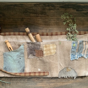 May include: A rustic, beige gardening apron with multiple pockets, each adorned with patches of varying colors and patterns. The apron features a plaid trim and holds wooden tools and dried herbs, suggesting a vintage or farmhouse aesthetic.