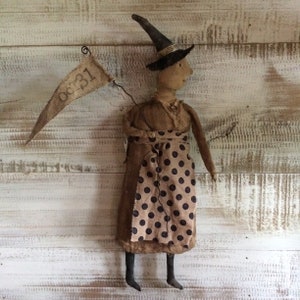 May include: A handmade rag doll witch wearing a black hat and a brown dress with black polka dots. She is holding a banner that says "Oct. 31".