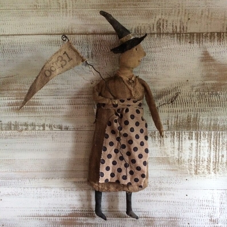 May include: A handmade rag doll witch wearing a black hat and a brown dress with black polka dots. She is holding a banner that says "Oct. 31".