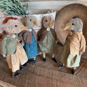 May include: Four handmade felt bird figurines dressed in vintage-inspired clothing. The birds are standing on a wooden surface. Each bird has a different colored dress and a button on its chest.