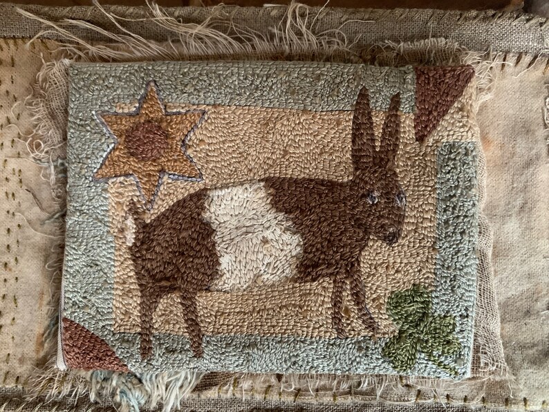 May include: A brown and white rug featuring a stylised image of a deer, a star, and a leaf. The rug is made from a textured fabric and has a rustic, vintage appearance.