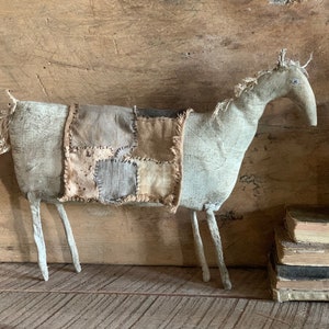 Op de afbeelding: Een handgemaakt primitief paardbeeldje van stof en patchwork. Het paard is grijs met een bruin en beige patchwork zadel en een lange, gescheurde staart.