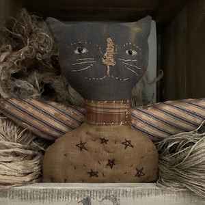 May include: A gray and brown fabric cat doll with wings, sitting in a wooden box with brown twine. The cat has a plaid collar and a brown fabric body with embroidered stars.