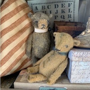 Puede incluir: Dos osos de peluche vintage, uno marrón y otro beige, sentados en una caja con una etiqueta vintage. Los osos tienen ojos de botón y llevan collares. El fondo incluye una almohada a rayas y una hoja de papel con el alfabeto impreso.