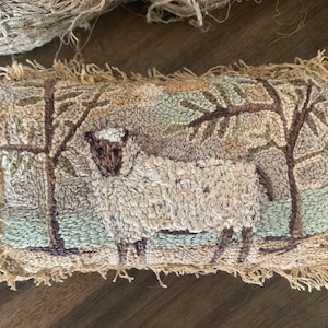 Op de afbeelding: Een klein, rechthoekig kussen met een geborduurd ontwerp van een schaap dat in een veld met bomen staat. Het kussen is bruin, beige en groen en heeft een rafelige, bruine rand.