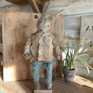 May include: A handmade fabric bunny rabbit wearing a brown jacket and blue plaid trousers. The bunny is standing on a wooden platform in front of a wooden background. There are yellow flowers in the background.