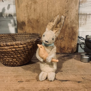 May include: A beige bunny rabbit toy with a blue ribbon around its neck is holding a small orange bird toy. The bunny is standing on a wooden surface.