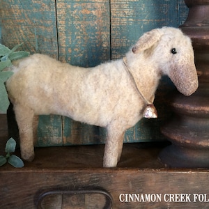 May include: A white, fluffy, handmade sheep figurine with a bell around its neck. The sheep is standing on a wooden surface with a blue and green painted background. The text "CINNAMON CREEK FOLK ART" is visible in the bottom right corner of the image.