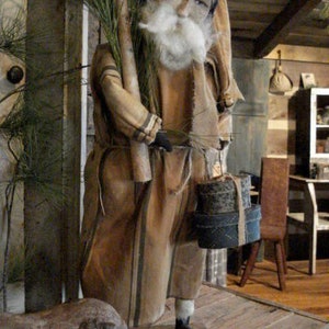 Op de afbeelding: Een handgemaakte primitieve stijl kerstmanpop met een lange witte baard, die een bruin-wit gestreepte robe en een zwarte hoed draagt. Hij houdt een bundel dennentakken en een stapel ingepakte cadeaus vast.