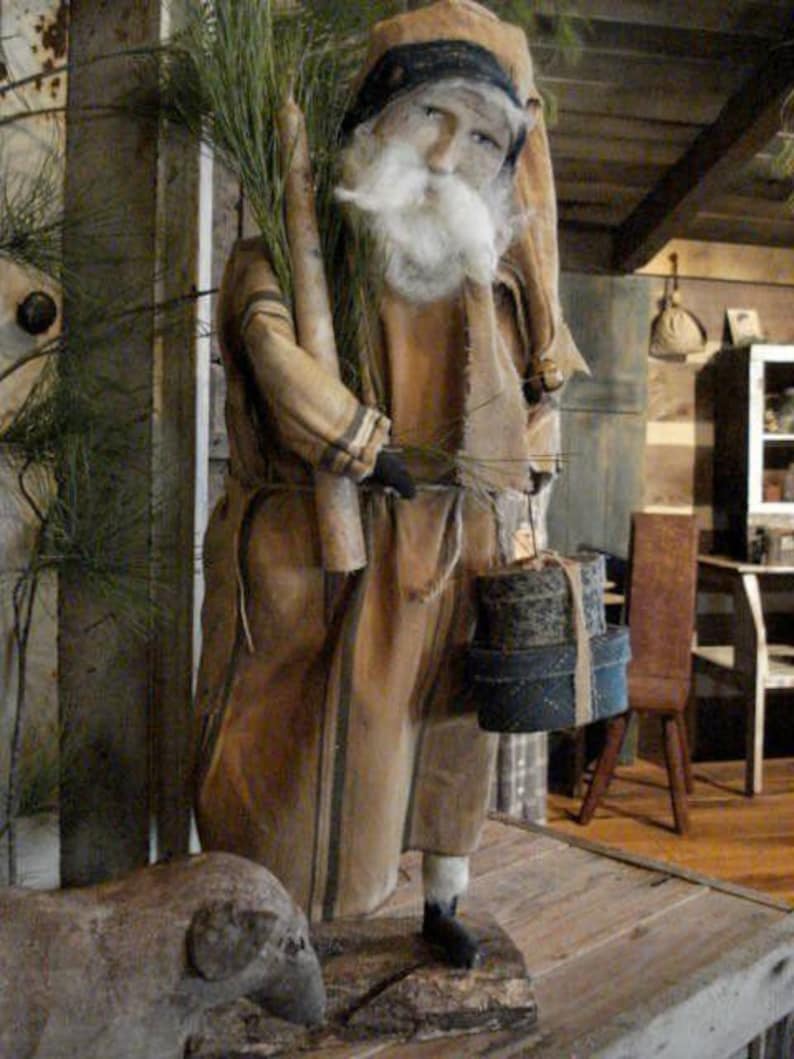 Op de afbeelding: Een handgemaakte primitieve stijl kerstmanpop met een lange witte baard, die een bruin-wit gestreepte robe en een zwarte hoed draagt. Hij houdt een bundel dennentakken en een stapel ingepakte cadeaus vast.