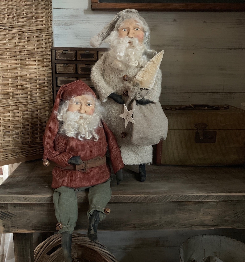 K&ouml;nnte beinhalten: Zwei handgefertigte Stoffpuppen, die als Weihnachtsmann verkleidet sind. Die Puppen sitzen auf einem Holztisch. Die Puppe links tr&auml;gt ein rot-gr&uuml;nes Outfit und einen wei&szlig;en Bart. Die Puppe rechts tr&auml;gt ein wei&szlig;es Outfit und einen wei&szlig;en Bart. Beide Puppen haben einen wei&szlig;en Bart und eine rote Nase.