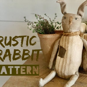 May include: A rustic, handmade rabbit doll with a striped fabric body and brown fabric ears. The doll is sitting on a wooden surface with a pot of green plants and a wooden bucket with straw and white eggs in the background. The text "RUSTIC RABBIT PATTERN" is written in brown letters on a white background.