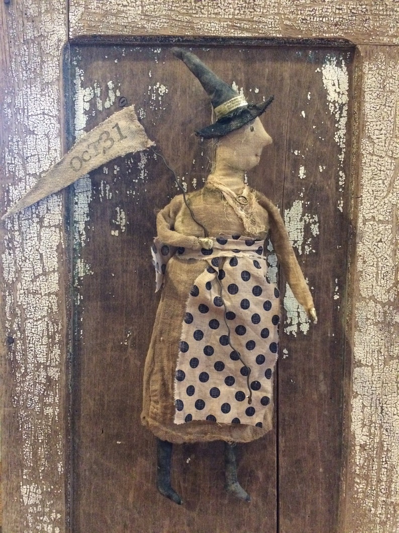 May include: A handmade rag doll witch wearing a black witch hat and a brown dress with black polka dots. The doll is holding a banner that says "Oct. 31".