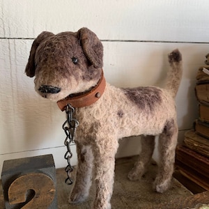 以下が含まれることがあります： 革の首輪と鎖が付いた茶色と灰色のフェルト製の犬のおもちゃ。犬は木製の表面に立っています。