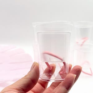 May include: A small, clear, cylindrical plastic cup containing a single pink elastic hair tie. The cup features a smooth, transparent wall and a flat base, with additional identical cups visible in the background, each holding a pink hair tie.
