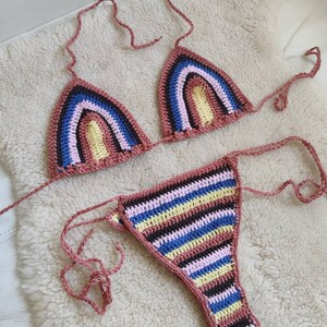 May include: A crocheted bikini top and bottom with a rainbow stripe pattern in pink, yellow, blue, brown, and black. The bikini is made with a thick yarn and has a simple design.